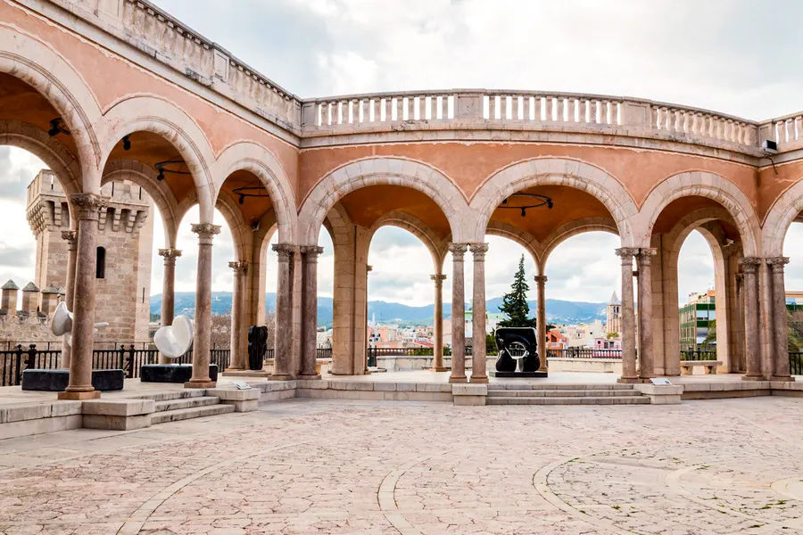 Offene Arkadenhalle mit rötlichen Rundbögen und Balustrade, dahinter die Stadt und die Berge im Dunst.