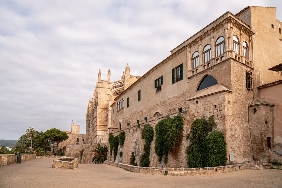 Der Palau de l'Almudaina in Palma von außen, ein langgestreckter Steinbau mit Zinnen, dahinter die Türme der Kathedrale.
