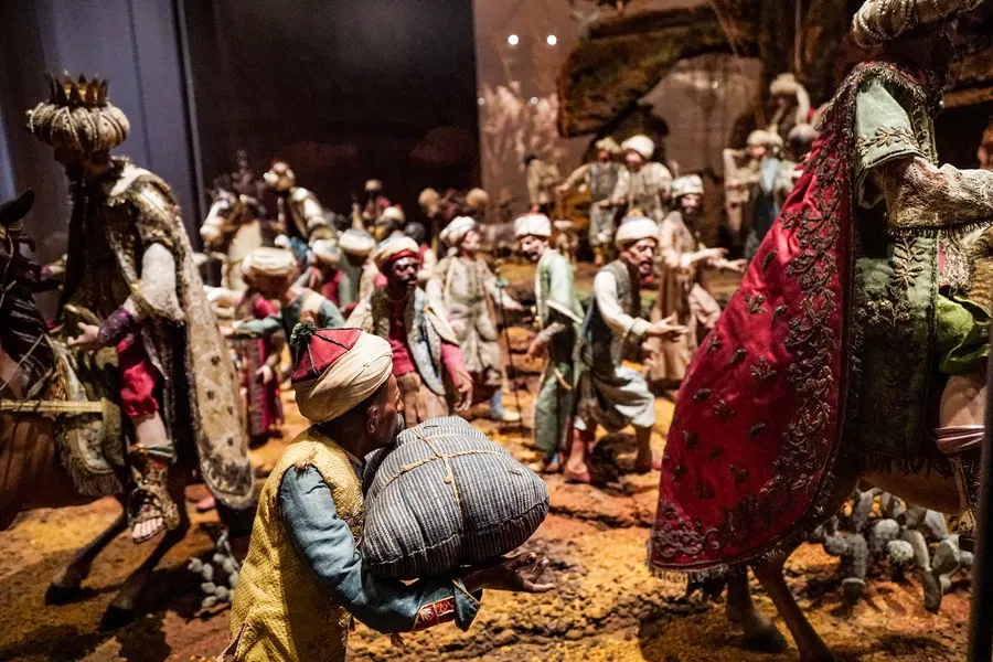 Ausschnitt aus der Krippe mit dem Zug der Könige: Figuren in Turbanen und bestickten Gewändern, ein Träger mit Bündel, dahinter Pferde unter rotem Samt.

