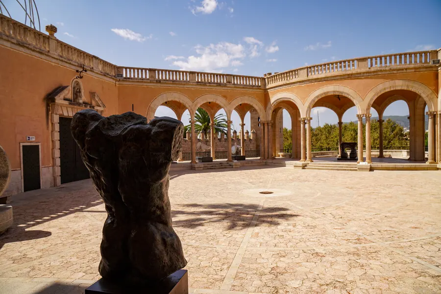 Weiter Blick über die gepflasterte Terrasse des Palau March mit umlaufender Arkade und Balustrade, im Vordergrund ein dunkler Bronzetorso auf schwarzem Sockel.