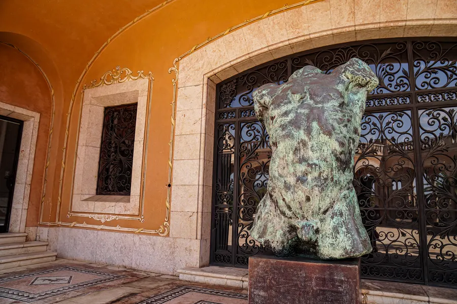 Ein grün patinierter Bronzetorso vor einem schmiedeeisernen Gitter und einer ockerfarbenen Wand mit Stuckrahmen, unten Mosaikboden.
