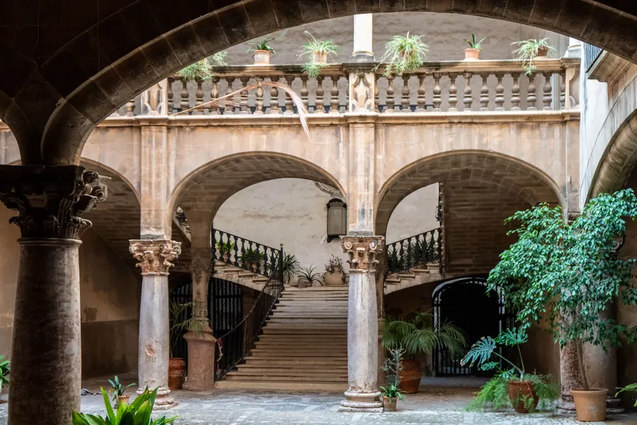 Innenhof des Stadtpalastes Can Vivot in Palma mit Arkaden auf schlanken Säulen, breiter Steintreppe und schmiedeeisernem Geländer.

