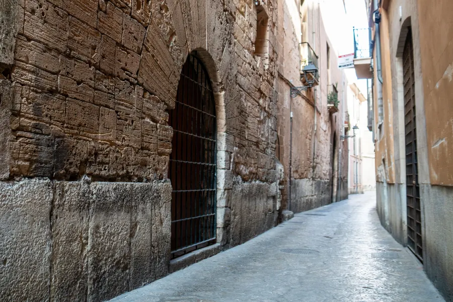 Enge Altstadtgasse in Palma mit einer alten Natursteinmauer links und einem vergitterten Fenster.
