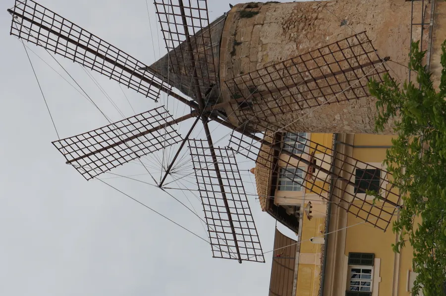 Eine der historischen Windmühlen von Es Jonquet in Palma von unten fotografiert, das hölzerne Flügelkreuz mit Gittersegeln vor bewölktem Himmel, daneben ein gelbes Wohnhaus.
