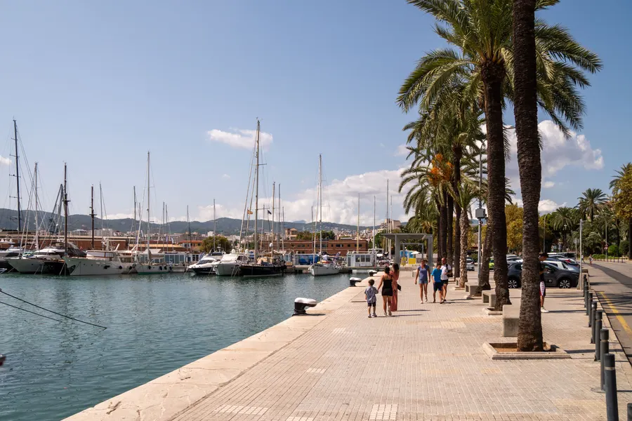 Breite Uferpromenade im Hafen von Palma mit Palmenreihe, Spaziergängern und Booten am Kai.