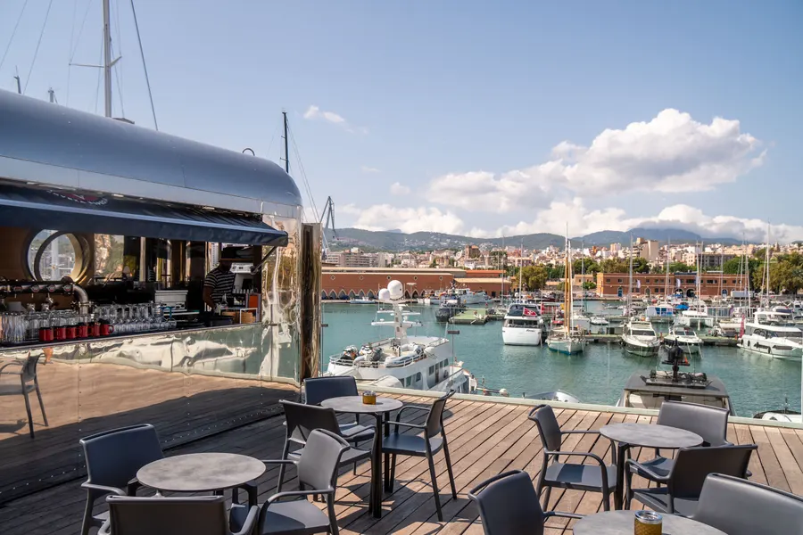 Terrasse eines Lokals am Hafen von Palma mit Tischen und Stühlen, dahinter Boote und die Stadt.