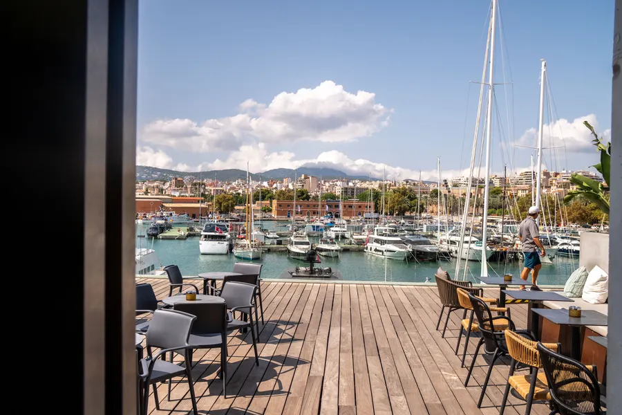 Holzdeck mit Bistrotischen direkt am Yachtbecken von Palma, dahinter Masten und die Stadt im Dunst.
