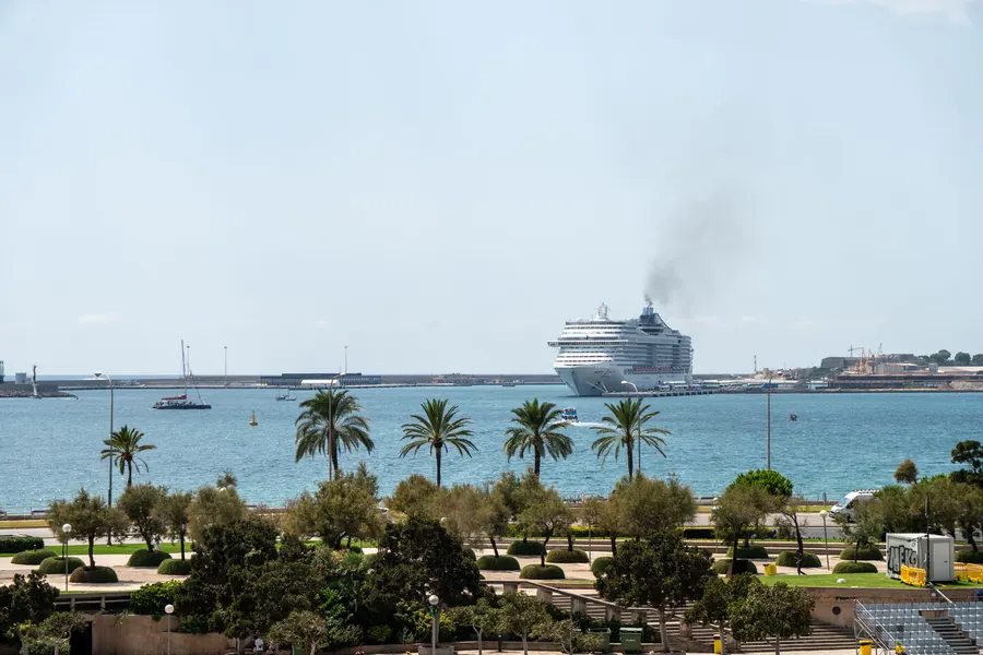 Ein Kreuzfahrtschiff verlässt den Hafen von Palma, im Vordergrund Palmen und die Grünanlage am Ufer.