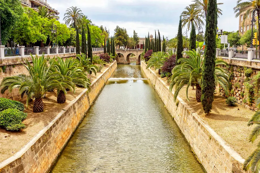 Der begradigte Wasserlauf der Riera in Palma zwischen gemauerten Ufern, Palmen und Zypressen.
