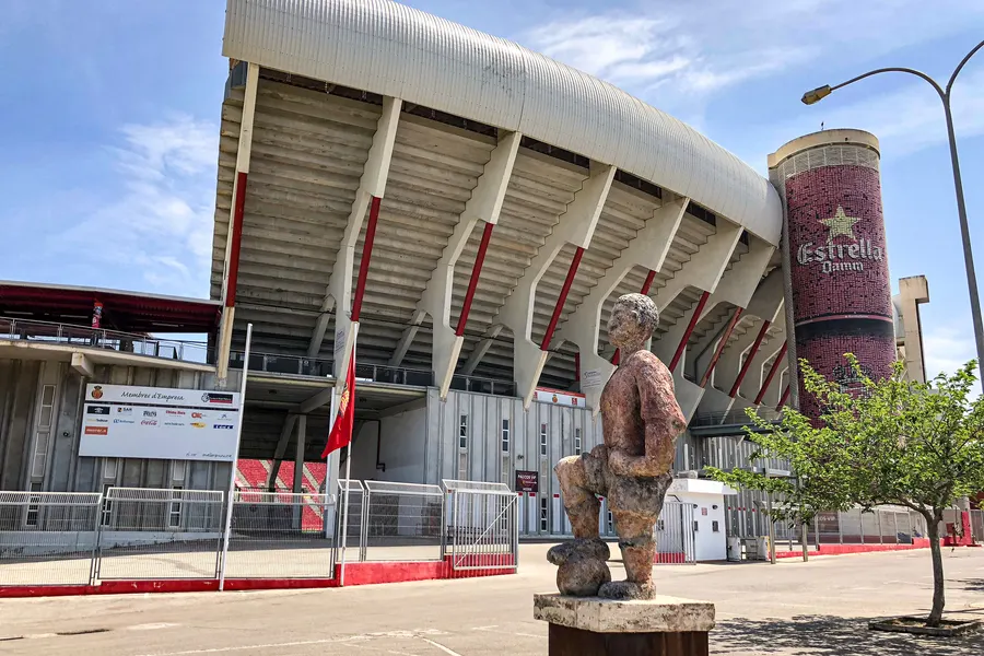 Das Estadi Mallorca Son Moix in Palma mit geschwungenem Dach über den Rängen, davor Bäume und die Zufahrt.
