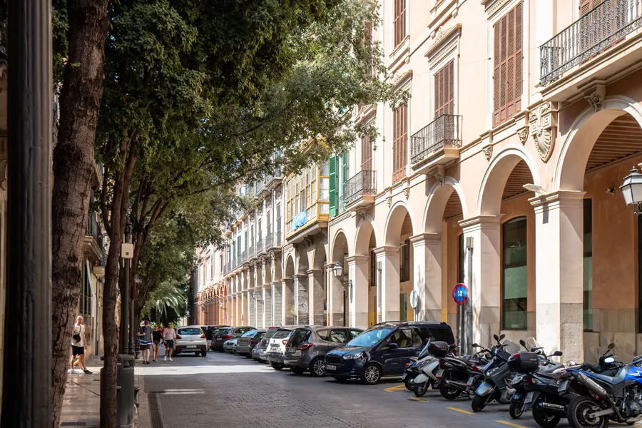 Straße in Palma mit langer Arkadenreihe, geparkten Motorrollern und Bäumen am Straßenrand.

