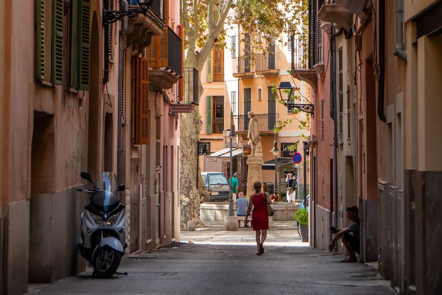 Schmale Gasse in Palma mit geparktem Motorroller, hohen Fassaden und Menschen am hellen Ende der Gasse.
