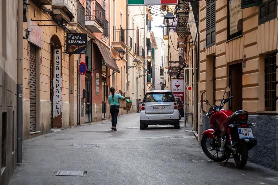 Gasse in Palma mit Restaurantschildern, geparkten Autos und einem Motorrad vor ockerfarbenen Häusern.
