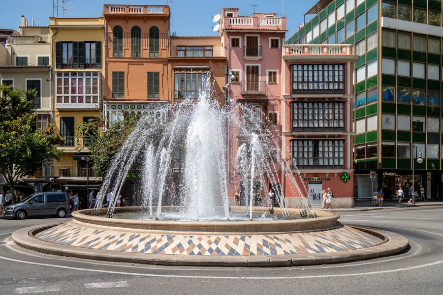 Der Springbrunnen auf der Plaça de la Reina in Palma, dahinter Häuser mit Erkerfenstern und ein Bürogebäude.
