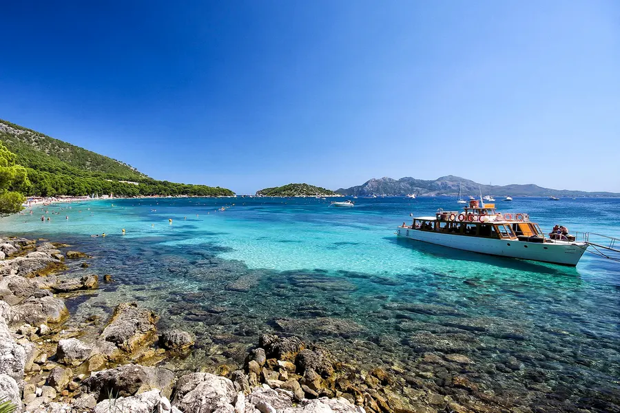 Felsiges Ufer am Rand der Bucht von Formentor mit klarem türkisfarbenem Wasser, davor ein weißes Ausflugsboot vor Anker, am Horizont eine Bergkette.