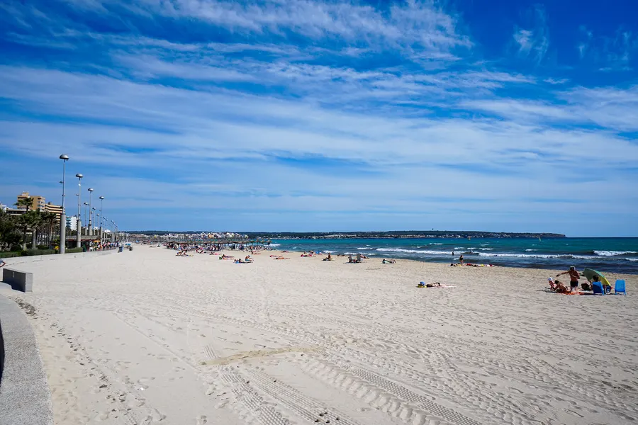 Gepflasterte Promenade mit Laternen und Bänken direkt neben dem breiten Sandstrand der Platja de Palma, dahinter Hotelbauten.
