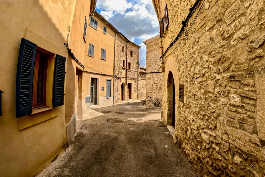 Enge, ansteigende Altstadtgasse mit ockerfarbenen und Natursteinfassaden, blauen Fensterläden und einer Straßenlaterne; am Ende lehnt ein Fahrrad an der Mauer, darüber ein Himmel mit weißen Wolken.