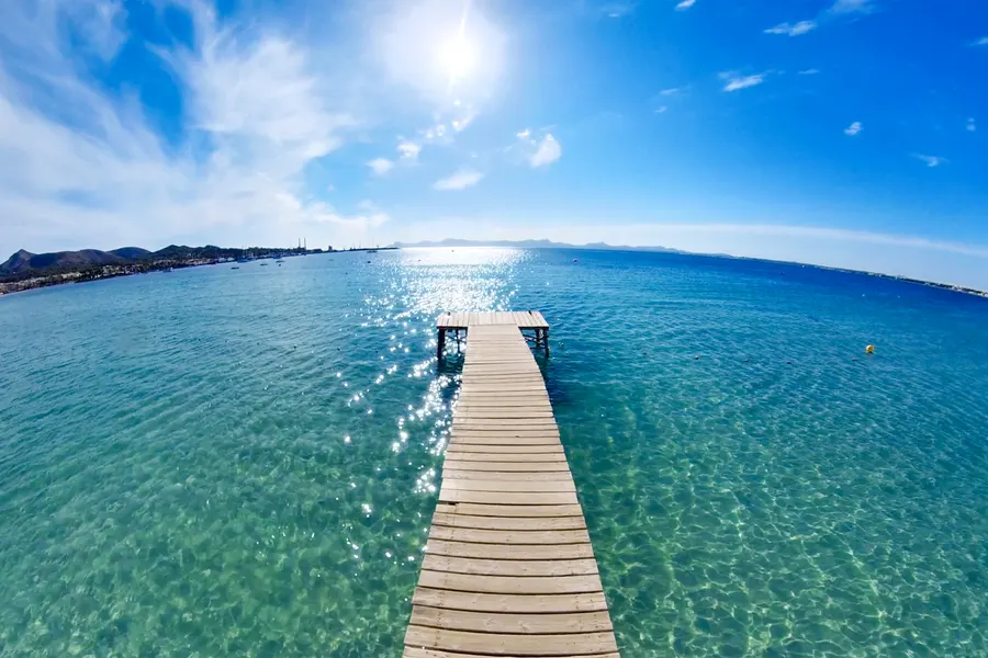 Hölzerner Badesteg, der geradewegs in kristallklares, türkis und blau schimmerndes Flachwasser der Bucht von Alcúdia führt; am linken Horizont die Bergkette der Halbinsel Victòria.