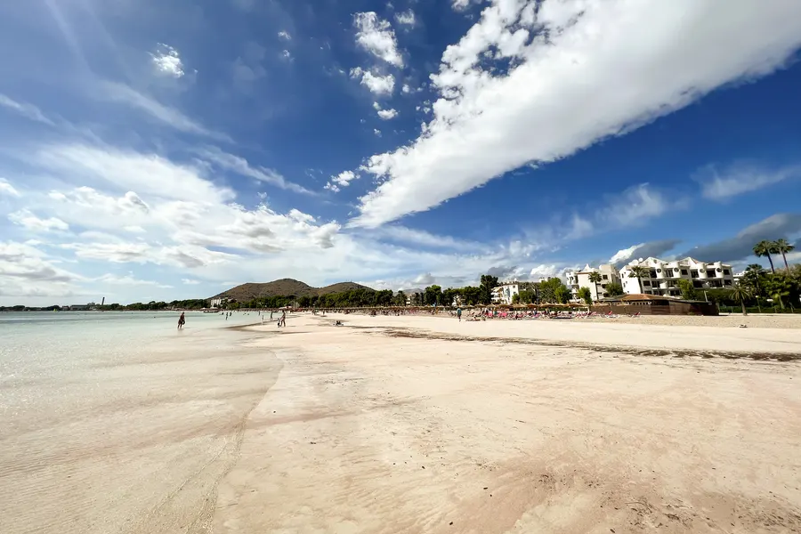 Weiter heller Sandstrand im September mit Reihen von Strohschirmen und Liegen vor Hotelbauten, links flach auslaufendes Wasser, am Horizont die Hügel der Halbinsel Victòria unter hohen Wolken.