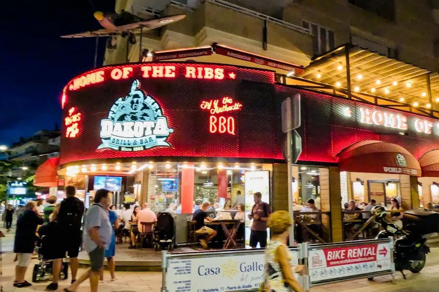 Abendlich beleuchtete Straßenecke mit rot leuchtender Leuchtreklame eines Grillrestaurants, gefüllte Terrassentische, Passanten mit Kinderwagen und ein Schild für Fahrradverleih.
