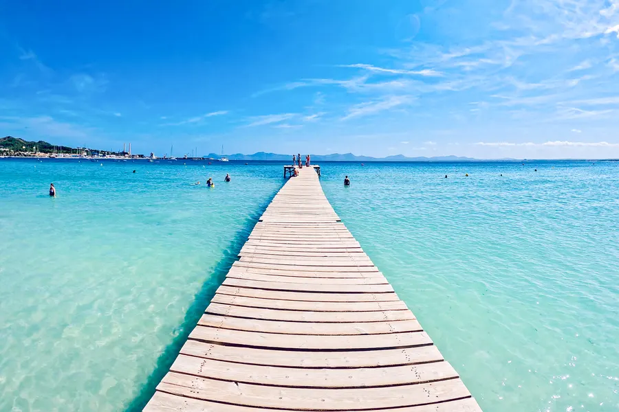 Langer hölzerner Badesteg, der vom Strand aus weit in das türkisfarbene, flache Wasser der Bucht von Alcúdia hinausführt, am Ende einzelne Badende.
