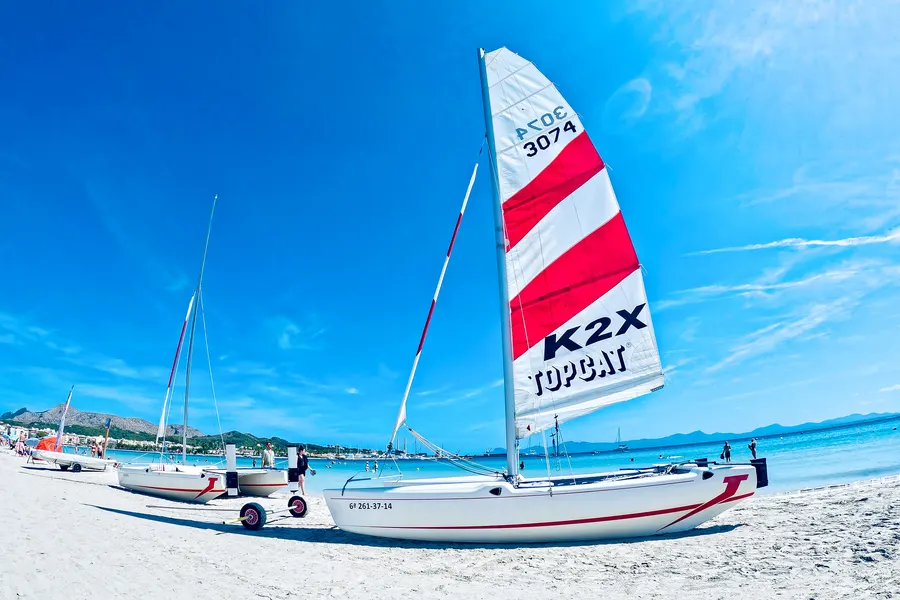 Aufgeriggte Katamarane mit rot-weißem Segel auf dem hellen Sand von Port d'Alcúdia, dahinter das türkisfarbene Wasser der Bucht.
