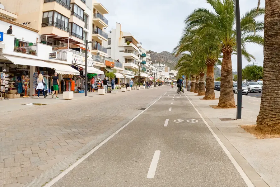 Breite Straße mit markiertem Radweg und Tempo-20-Markierung, links Läden mit Markisen und Auslagen unter Wohnhäusern, rechts eine Reihe hoher Dattelpalmen, im Hintergrund ein Bergrücken.
