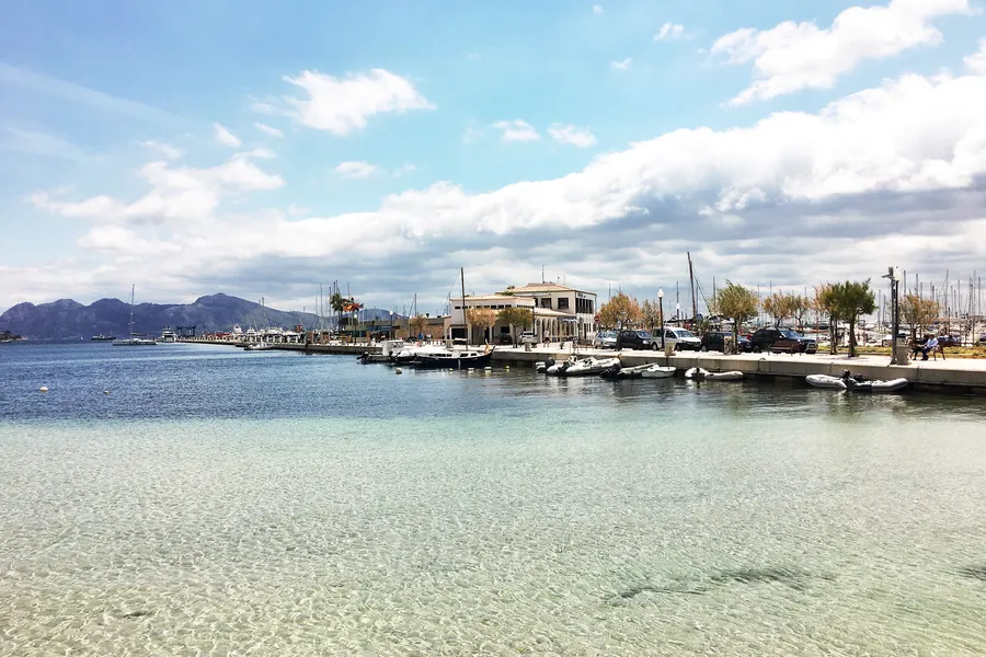 Blick über das glasklare Flachwasser der Bucht auf den Hafen von Port de Pollença mit Booten, Palmen und der niedrigen Uferbebauung, dahinter die Berge der Tramuntana.