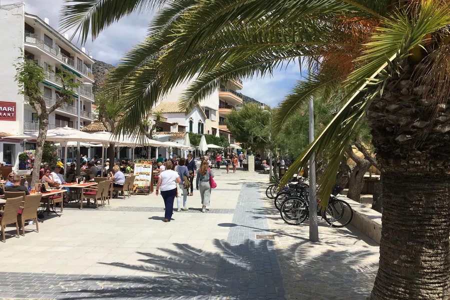 Belebte Uferpromenade von Port de Pollença mit Straßencafés unter Sonnenschirmen, Palmen, Radfahrern und Passanten.