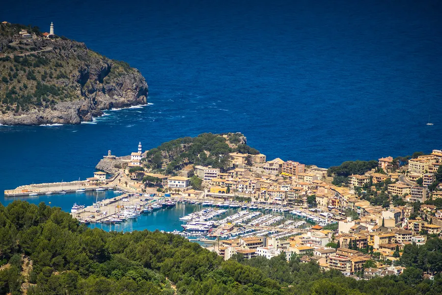 Ockerfarbene und helle Häuserzeile am Ufer von Port de Sóller, davor Segelboote im türkisen Wasser und die Promenade.