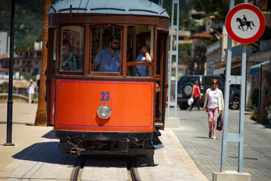 Fahrgäste, die sich aus den offenen Fenstern eines Wagens der Straßenbahn von Sóller lehnen, daneben Schilf und Gleis.
