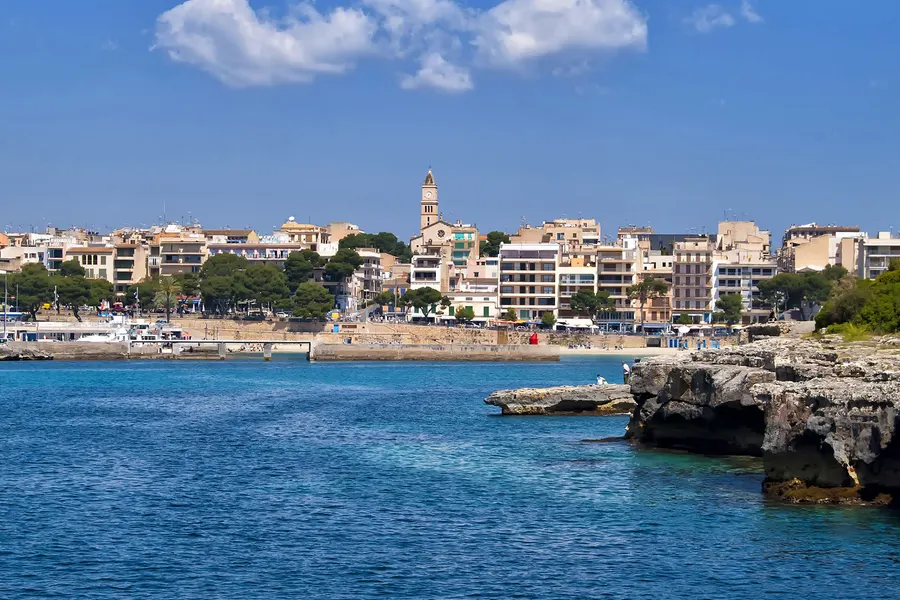 Blick über das Wasser auf die Hafeneinfahrt von Porto Cristo, links die felsige Küste, dahinter die Bebauung des Orts.

