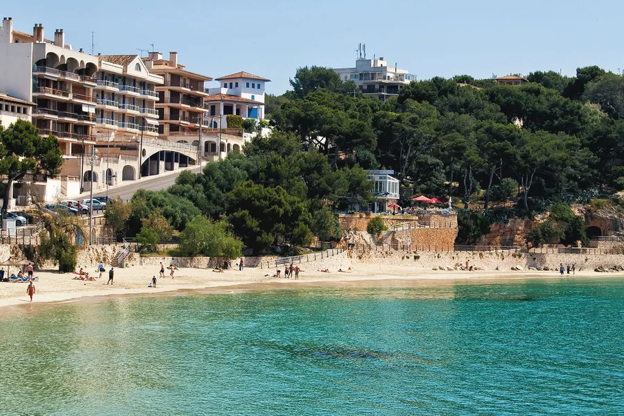 Felsige, bewachsene Küste am Rand der Bucht von Porto Cristo mit klarem Wasser und einzelnen Gebäuden auf der Kante.
