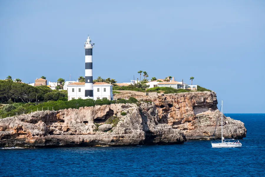 Der schwarz-weiß geringelte Leuchtturm von Portocolom auf der Felskuppe über der Hafeneinfahrt, davor das offene Meer.

