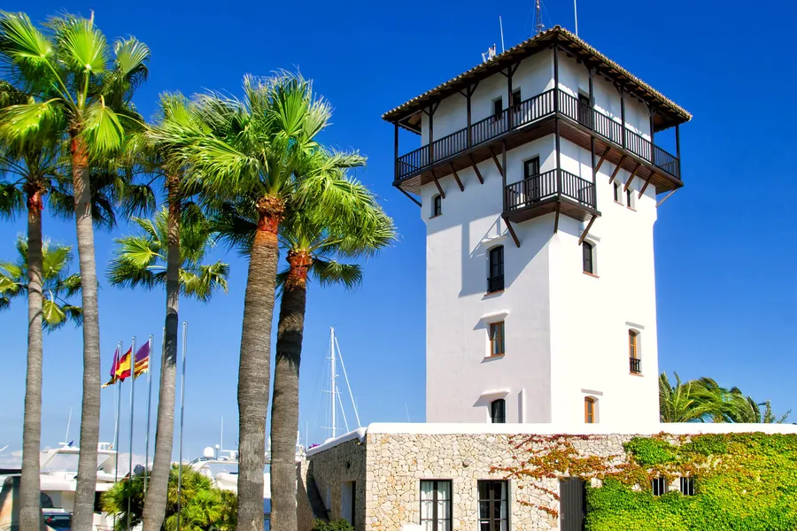 Weißer quadratischer Turm mit hölzerner Loggia und Ziegeldach am Hafen von Puerto Portals, davor hohe Palmen und Fahnenmasten.