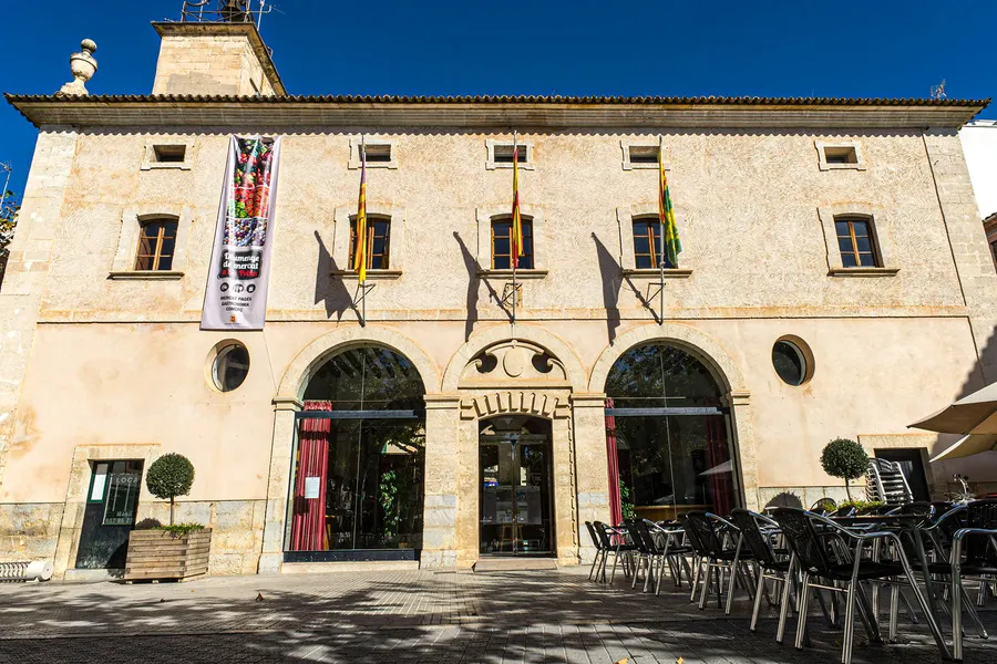 Langgestrecktes Gebäude aus hellem Sandstein in sa Pobla mit drei großen Rundbögen im Erdgeschoss, Fahnen über dem Eingang und Caféstühlen davor.