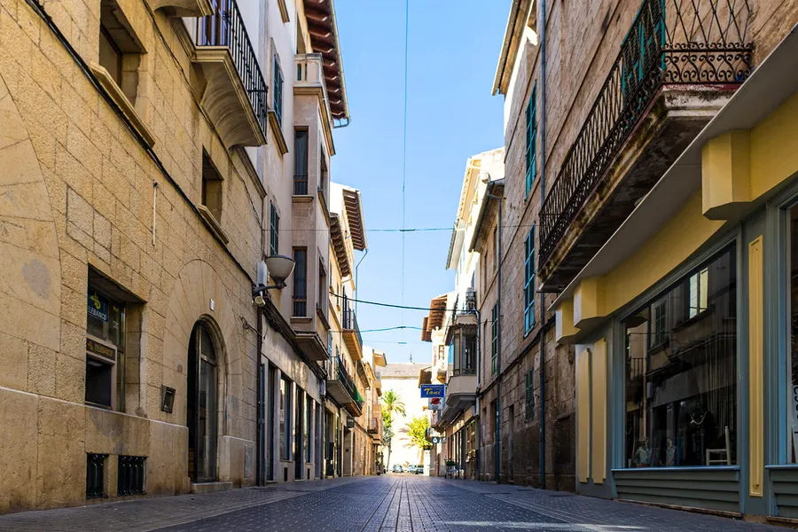 Enge Geschäftsstraße in sa Pobla mit Sandsteinfassaden, schmiedeeisernen Balkonen und Ladenfronten im Erdgeschoss.
