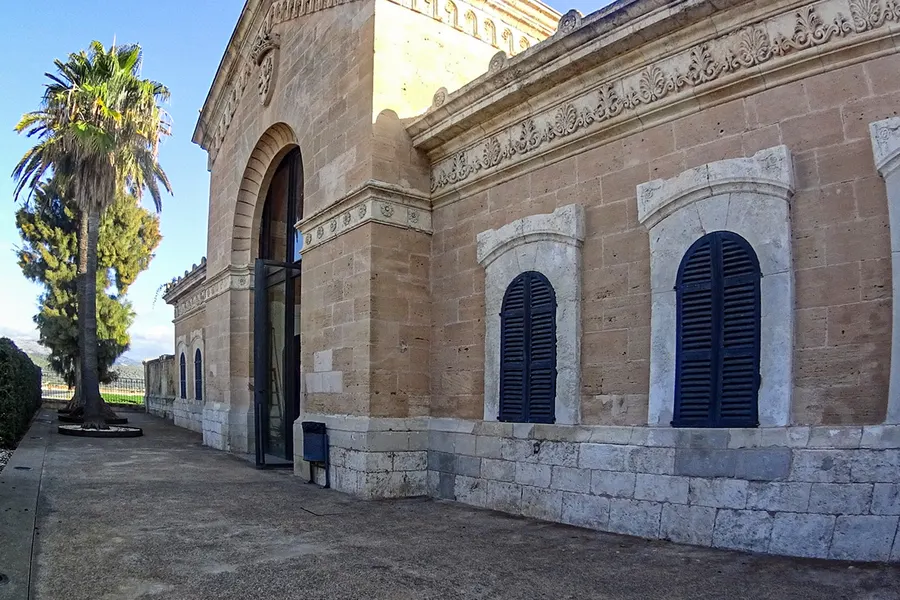 Öffentliches Gebäude in sa Pobla aus hellem Sandstein mit Bogenfenstern, dunkelblauen Läden und verziertem Gesims, daneben eine Palme.
