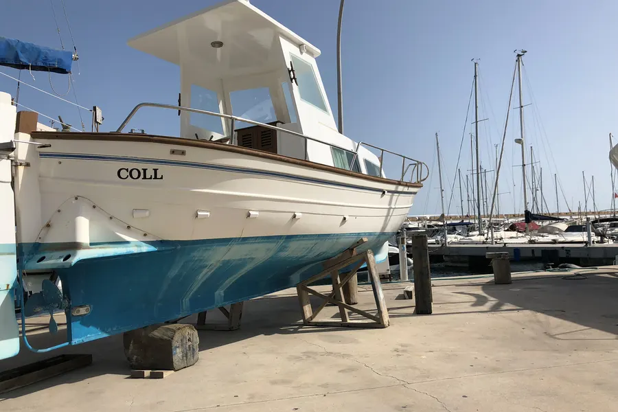 Weißblaues Fischerboot auf Böcken an Land im Hafen von Sa Ràpita, dahinter Masten und Stege.
