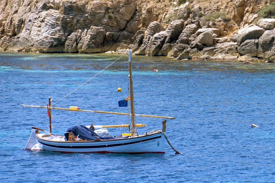 Kleines hölzernes Segelboot im tiefblauen Wasser vor der hellen Felswand der Dragonera.
