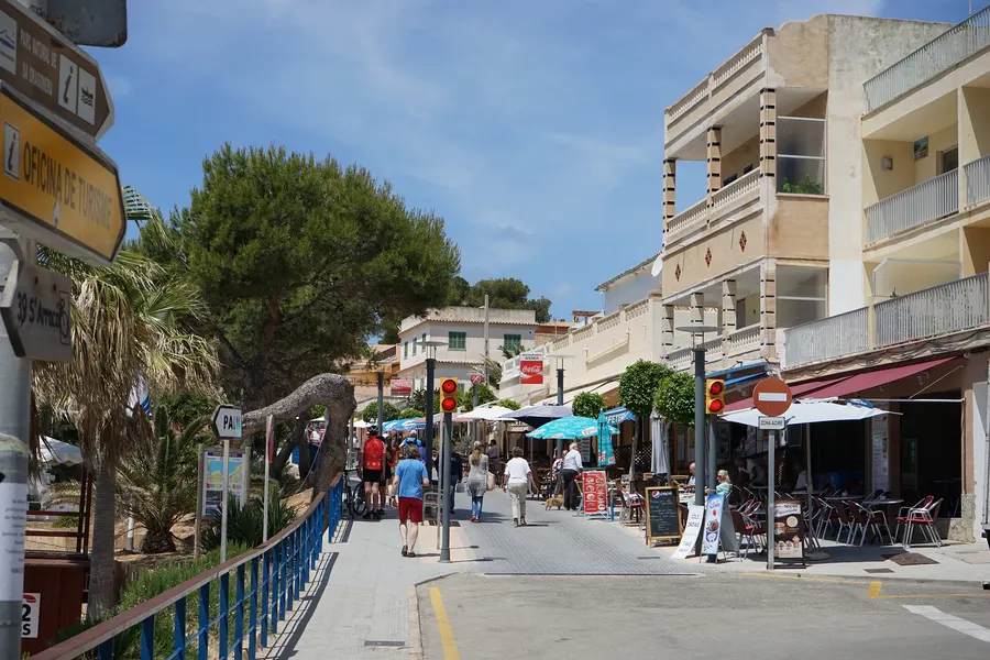 Hauptstraße von Sant Elm mit Wegweisern, Terrassen unter Sonnenschirmen und einem mehrstöckigen Gebäude, davor Kiefern.
