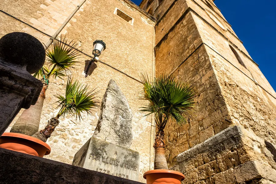 Aufsicht auf die massive Sandsteinmauer der Kirche von Sant Joan mit einem alten Gedenkstein und Kübelpalmen davor.
