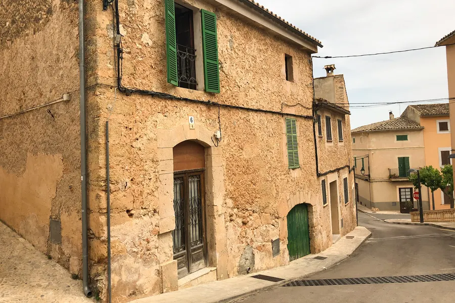 Ansteigende Gasse in Santa Eugènia zwischen alten Natursteinhäusern mit grünen Läden und Balkonen.
