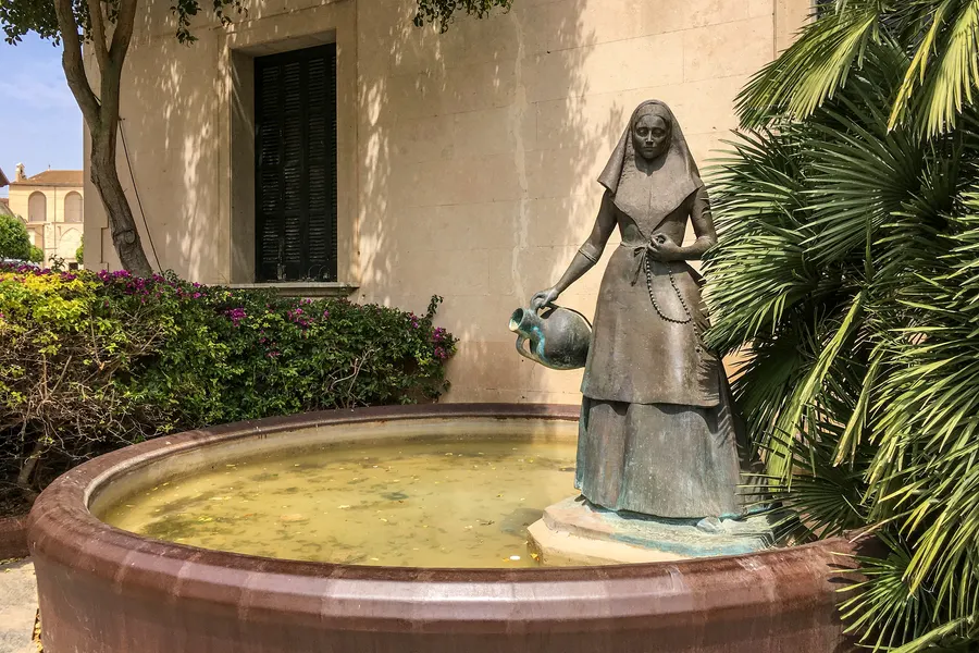 Bronzefigur einer Bäuerin mit Korb auf einem runden Brunnen in Santa Margalida, dahinter eine Palme und die Kirchenmauer.