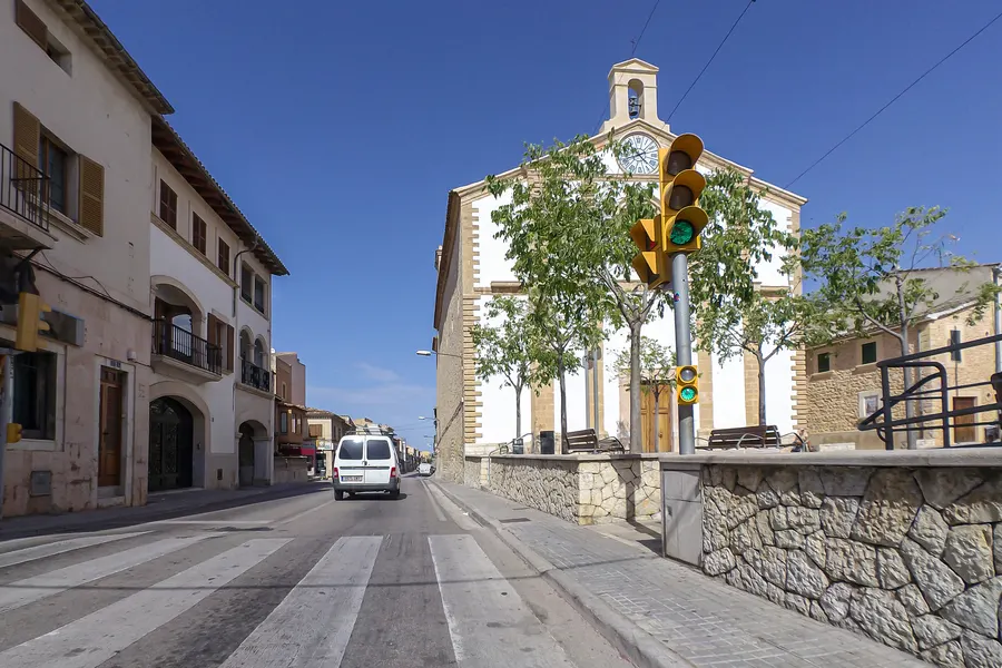 Heller Klosterbau mit Glockengiebel und Uhr an der Durchgangsstraße von Santa Maria del Camí, davor Bäume und eine Ampel.