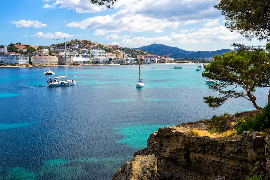 Zwei ankernde Segelboote in der türkisen Bucht von Santa Ponça, im Hintergrund die Bebauung des Orts und die Berge.
