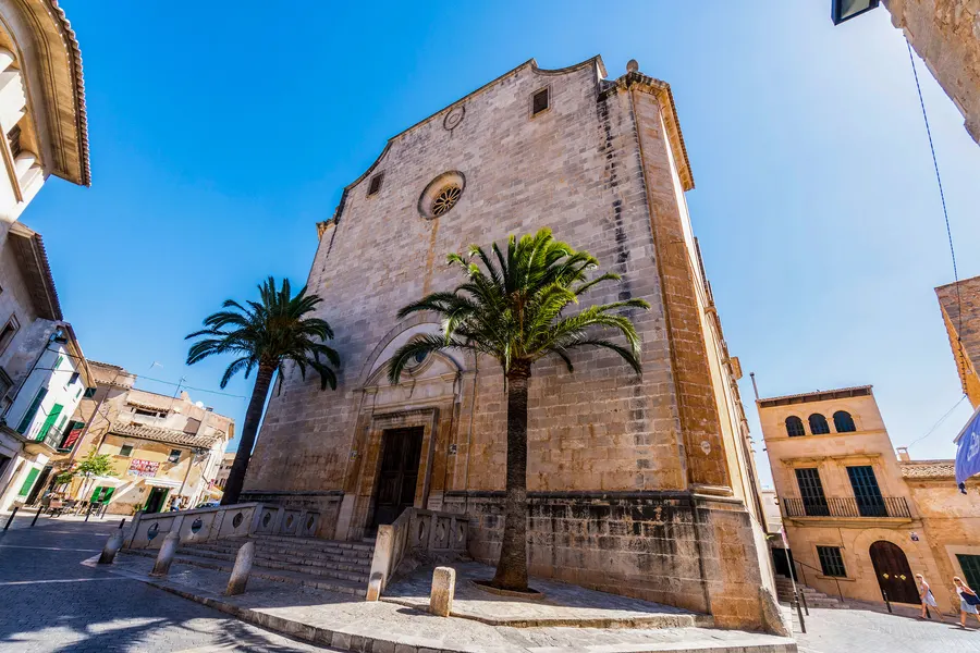Die wuchtige Sandsteinfassade der Kirche Sant Andreu in Santanyí mit Rundfenster und Portal, davor zwei Palmen und die Häuser der Plaça unter blauem Himmel.