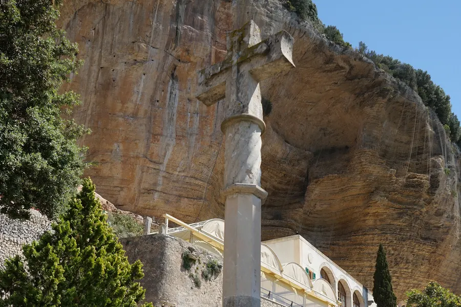 Steinernes Kreuz auf einer hohen Säule vor der überhängenden Felswand am Kloster Gràcia, daneben Zypressen und Bäume.