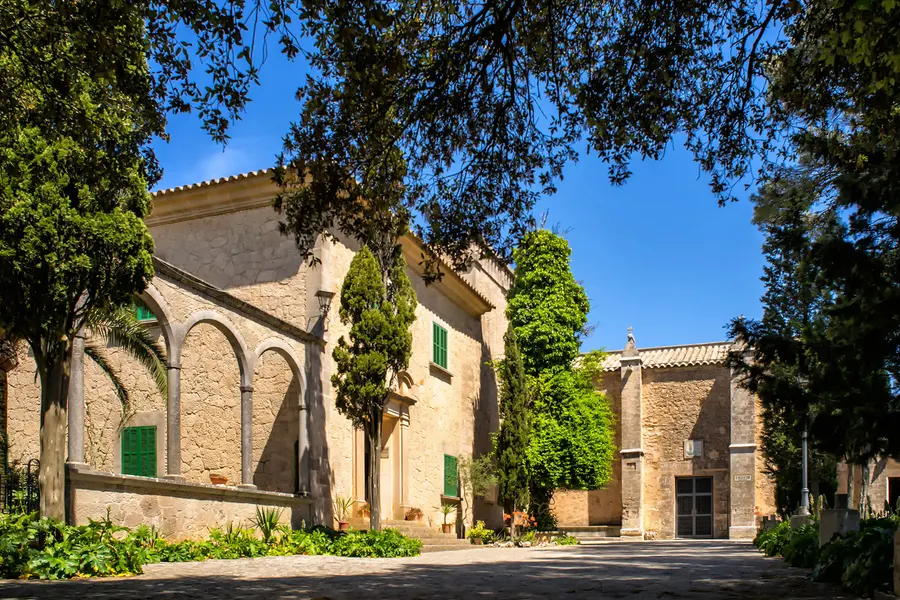 Innenhof des Klostergeländes Cura mit Bogengang, Steinhäusern, Zypressen und Kletterpflanzen unter blauem Himmel.
