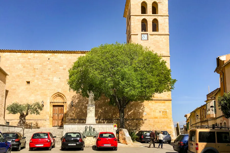 Die Pfarrkirche von Sencelles aus hellem Sandstein mit hohem Glockenturm, davor ein Platz mit Baum, Denkmal und parkenden Autos.
